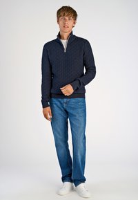 Navy gebreide trui met een gestructureerd ruitpatroon en een halve rits, gecombineerd met lichtblauwe rechttoe rechtaan spijkerbroek en witte sneakers.