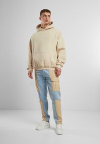 Beige Übergröße Hoodie mit Fronttasche, kombiniert mit hellblauen Cargo-Hosen mit beigen Stoffakzenten und mehreren Taschen, getragen mit weißen Sneakern.