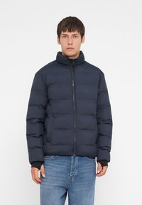 Marineblå puffjakke med høy krage, horisontal quiltet tekstur, glidelås foran, og to sidelommer. Båret over lyseblå jeans.