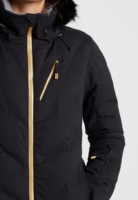 Veste noire isolante avec capuche bordée de fourrure, fermeture éclair dorée et plusieurs poches zippées pour le rangement. Le tissu texturé ajoute de la dimension.