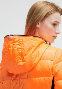 Puffer jacket in bright orange nylon with a hood, featuring quilted texture and a zippered front closure, showing the logo tag near the collar.
