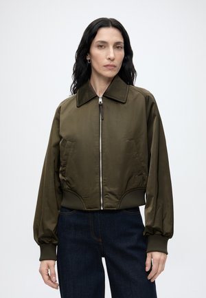 Kvinde med langt mørkt hår iført olivengrøn lynlåstrøje og mørkeblå jeans, stående mod en ensfarvet lys baggrund.