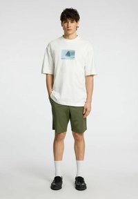 Vit t-shirt med kort ärm och en blå grafik, tillsammans med gröna shorts. Modellen bär svarta slip-on skor och vita strumpor.