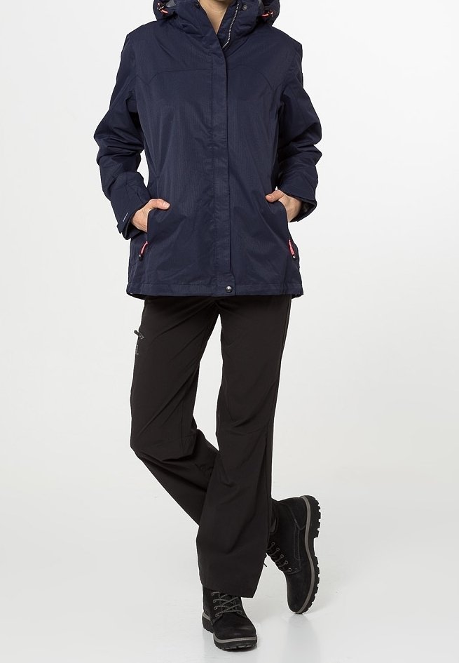 Personne portant une veste à capuche bleu marine, un pantalon noir et des bottes noires, debout les mains dans les poches devant un fond uni.
