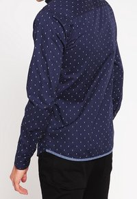 Marineblauw langemouwen shirt met een klein wit bladerenpatroon, knop-down kraag en een gebogen zoom. De stof lijkt soepel met een casual pasvorm.