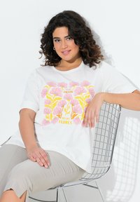 T-shirt blanche à manches courtes avec un motif floral rose et jaune et le texte "Fiesta de las Flores." Pantalons gris texturés.