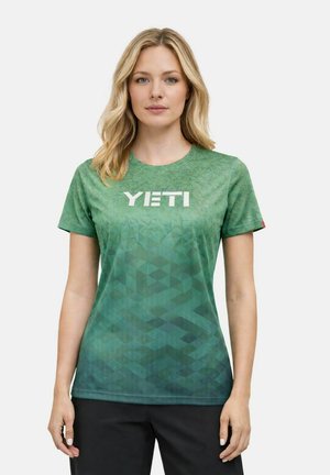 Frau mit blondem Haar, die ein grünes YETI-T-Shirt mit geometrischem Muster und schwarze Hose trägt, vor einem schlichten Hintergrund stehend.