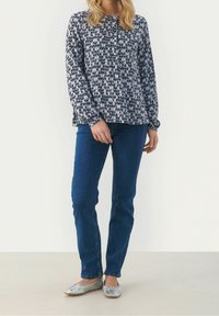Blusa a maniche lunghe con un motivo geometrico blu navy e bianco, con bottoni frontali e vestibilità comoda. Abbinata a jeans blu scuro e ballerine argentate.