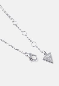 Zilveren ketting met een driehoekige hanger waarin "GUESS" gegraveerd staat, en een karabijnsluiting. Lichtgewicht en gestructureerde schakels.