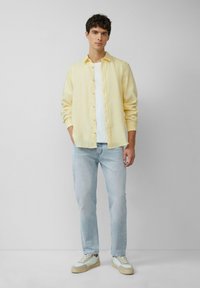 Chemise bouton en jaune clair sur un T-shirt blanc, associée à un jean bleu clair et des baskets claires, se tenant devant un fond neutre.