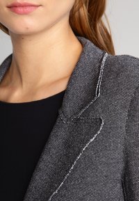 Blazer gris con una tela texturizada, solapas con muesca y acentos de costura blanca. Llevado sobre una blusa negra, mostrando el detalle del cuello.