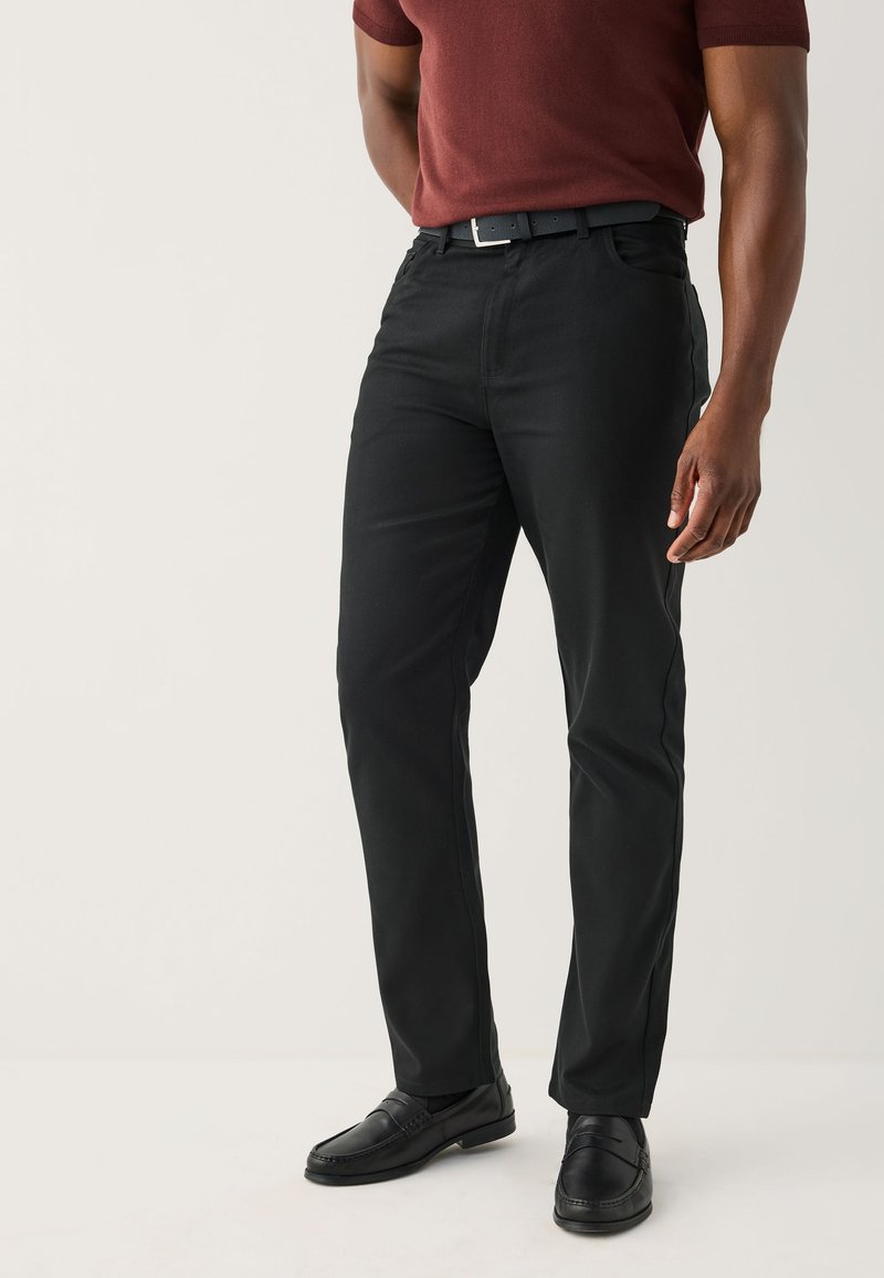 Homme portant un pantalon noir, une ceinture noire, des mocassins noirs et une chemise à manches courtes bordeaux, debout devant un fond clair uni.