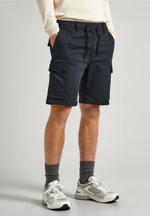Shorts cargo bleu marine en mélange de coton, avec deux poches latérales et une coupe ample. Associés à des baskets claires et des chaussettes grises.