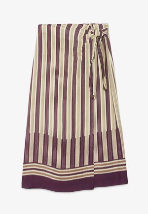 Vrouw staand, draagt een witte blouse met ruches, een gestreepte midi-rok met een strik aan de zijkant en witte sandalen met hakken op een betonnen vloer.