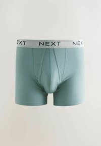 Világoskék férfi boxer alsónadrág fehér elasztikus derékszíjjal, amelyen fekete betűkkel a "NEXT" felirat található, egy sima világos háttéren.