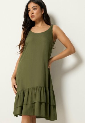 Femme aux longs cheveux foncés portant une robe sans manches vert olive avec un ourlet à volants double couche, debout avec une main sur la hanche.