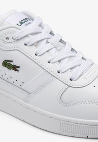 Sneaker in pelle bianca con accenti texturizzati, lacci bianchi piatti, piccolo logo di coccodrillo verde e dettagli perforati lungo il lato.