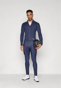 Tamsiai mėlynas dviračių kostiumas, kuriame yra užsegamas ilgomis rankovėmis viršus ir aptemptos tamprės, pagaminti iš lygaus, tampraus audinio. Komplekte yra šalmas.