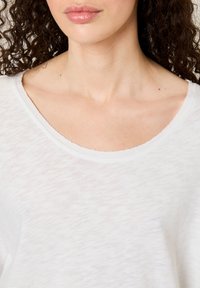 Naine lokkis tumepruuni juustega, kellel on seljas valge sügava kaelusega särk, mis paljastab kaela ja rangluu ala.