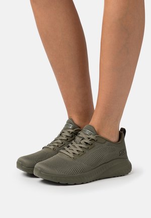 Baskets tricotées vert olive avec lacets portées aux pieds, dotées de semelles rembourrées et d'un logo discret de la marque sur le côté.