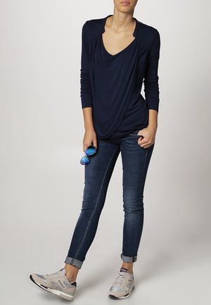 Top azul marino de manga larga con frente drapeado, jeans ajustados oscuros y zapatillas claras. Sosteniendo gafas de sol azules, de pie sobre un fondo neutro.