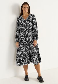 Schwarzes Midi-Kleid mit einem weißen Blumenmuster, langen Ärmeln, V-Ausschnitt und lockerer Passform. Schwarze Schuhe ergänzen das Outfit.