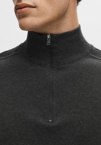 Tmavě šedý svetr na zip s vysokým límcem, vyrobený z texturované pleteniny, který má malý stříbrný zip a minimalistický design.