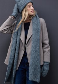 Abrigo gris de mezcla de lana, gorro de punto color teal, bufanda y guantes. La bufanda es larga y esponjosa; los guantes son texturizados y ajustados. Jeans azules. Fondo neutro.