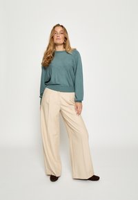 Mēteļu krāsas adīta džemperis ar garām piedurknēm un pieguļošu apakšmalu, savienots ar augsto jostasvietu, platajiem beidža krāsas biksēm un tumši brūniem kurpēm.