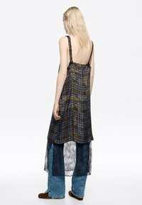 Vista trasera de un vestido de capas con una parte superior de cuadros marrones, tirantes de encaje y una sobrefalda de encaje negro, usado sobre jeans de pierna ancha.