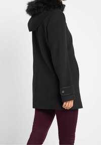 Svart huva över rock med fuskpälsdetaljer, har sidofickor och knäppta ärmslut. Slät textur, skräddarsydd design och knälång.