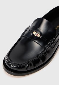 Μαύρο δερμάτινο λουστρίνι με γυαλιστερή επιφάνεια, που διαθέτει ραμμένο σχέδιο, διακοσμητική ταινία και μικρή χρυσή ετικέτα μπροστά.