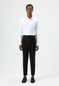 Chemise blanche moulante à manches longues avec boutons, associée à un pantalon noir plissé taille haute et des mocassins foncés. Tissu lisse, design ajusté.