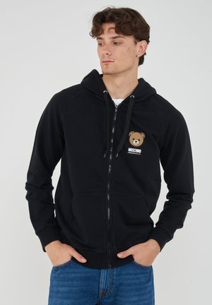 Giovane uomo che indossa una felpa nera con zip con logo dell'orsetto e scritta "Moschino Underbear", mani nelle tasche, guarda di lato.