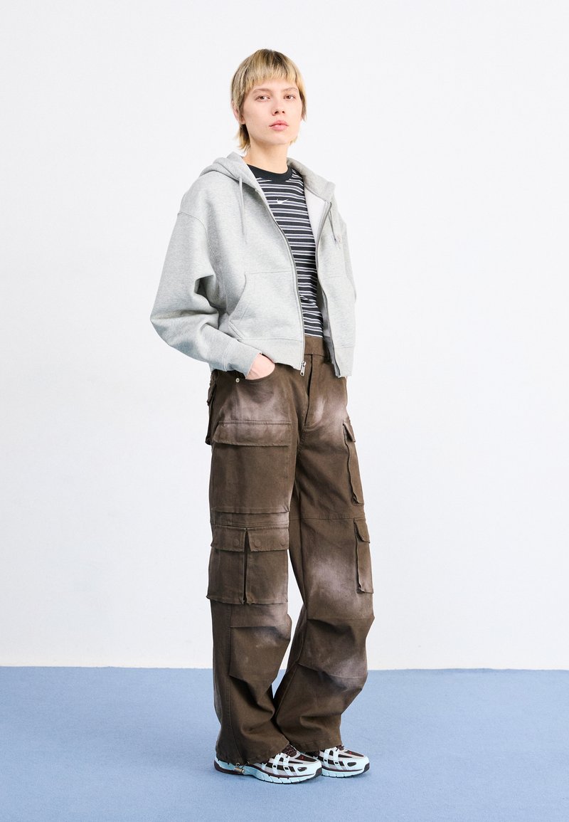 Veste à capuche zippée grise, chemise rayée noire et blanche en dessous, pantalon cargo brun avec plusieurs poches, et chaussures de sport bleu clair.