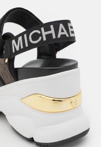 Sandale plateforme noir et blanc avec accent doré et une sangle noire de marque présentant un texte blanc partiellement visible "MICHAE".