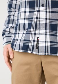 Camisa de cuadros en navy, gris y blanco con cierre de botones. Presenta una etiqueta en el dobladillo, combinada con pantalones caqui. Tejido texturizado.