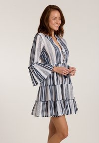 Blau-weiß gemustertes Kleid mit Glockenärmel und einem gestuften Rüschen-Saum. Mit geometrischen Designs und einem V-Ausschnitt. Leichtes Material.