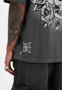 Camiseta gris de manga corta con un gráfico de cráneo y rosa, con textura desgastada y un detalle de logo impreso en el dobladillo.