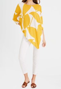 Gelbes und weißes Poncho mit einem Blattmuster, das einen asymmetrischen Saum aufweist. Kombiniert mit weißen eng anliegenden Hosen und braunen Rutschsandalen.