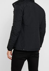 Černá izolovaná bunda z nylonu s vysokým límcem, bočními kapsami a prošitými detaily na rukávech.