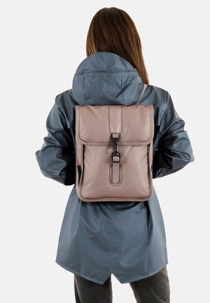 Zaino in pelle rosa con chiusura a pattina, spallacci regolabili e un dettaglio della chiusura frontale, indossato sopra una giacca impermeabile blu.