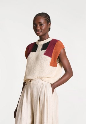 Jeune femme aux cheveux tressés portant un haut sans manches en maille avec des blocs noirs, bordeaux et orange, et un pantalon taille haute de couleur claire.