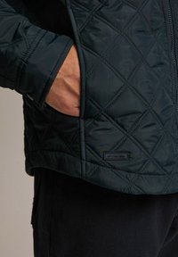 Pikowana ciemnozielona kurtka z zamkiem błyskawicznym i bocznymi kieszeniami, cechująca się teksturowaną powierzchnią i małą metką z logo u dołu.