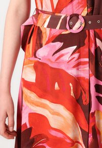Robe florale avec des motifs vibrants de rouge, rose et orange ; ceinturée à la taille avec une boucle circulaire ; texture de tissu lisse.