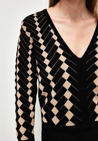 Pull noir et beige à motifs, avec un col en V, un design géométrique en forme de losange et un tissu en tricot texturé. Les couleurs à fort contraste mettent en valeur le motif.