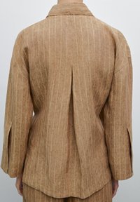 Veste en lin marron à rayures verticales, avec un pli au dos, des manches larges et un col. La texture semble légèrement froissée.