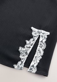 Tessuto nero con una texture a coste caratterizzato da un bordo in pizzo bianco e un motivo floreale lungo una spaccatura su un lato.