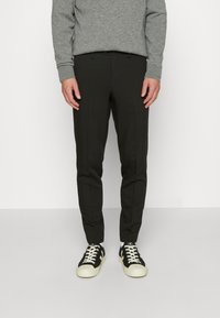 Svarta avsmalnande byxor med en slät yta, som har sidofickor och bältesöglor, kombinerat med svartvita sneakers. Grå tröja ovanför.