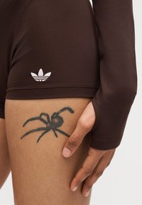 Χέρι που ακουμπάει στον μηρό δείχνοντας ένα μαύρο τατουάζ αράχνης, φοράει σκούρα καφέ σορτς Adidas με λευκό λογότυπο Adidas.